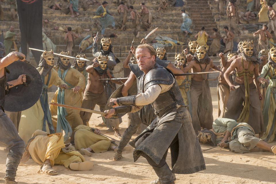 Iain Glen as Jorah Mormont fighting masked enemies in a gladiatorial arena.