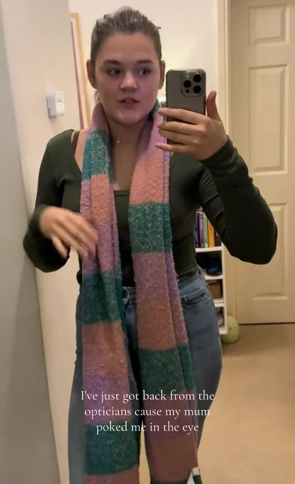 A young woman wearing a long-sleeved shirt, jeans, and a pink and green striped scarf, records herself in a mirror.