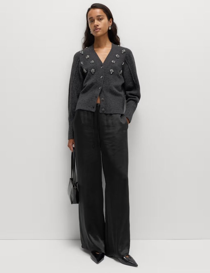 A woman wearing a gray cardigan with embellishments and black satin wide-leg trousers.
