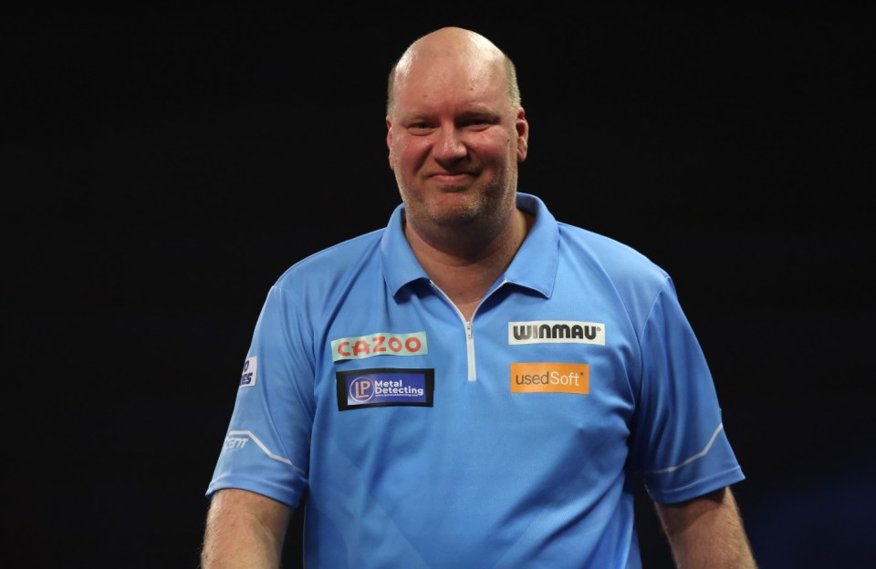 A male darts player in a light blue shirt with several sponsors smiles at the camera.