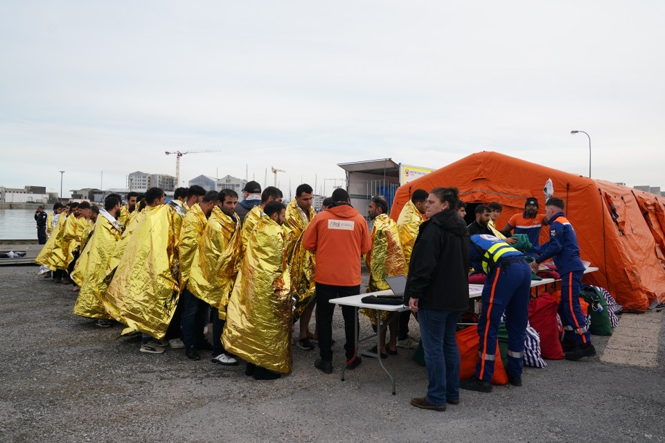 Migrants wrapped in golden thermal blankets lining up, with aid workers and an orange tent nearby.