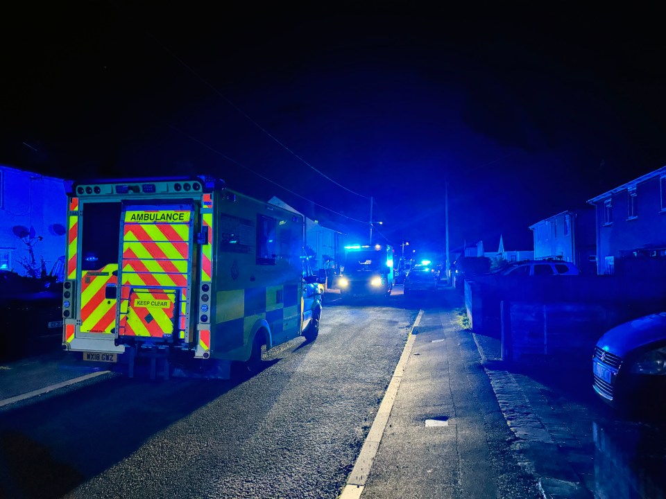 An ambulance with its lights on at night, on a street lined with residential houses and other emergency vehicles with flashing lights in the distance.