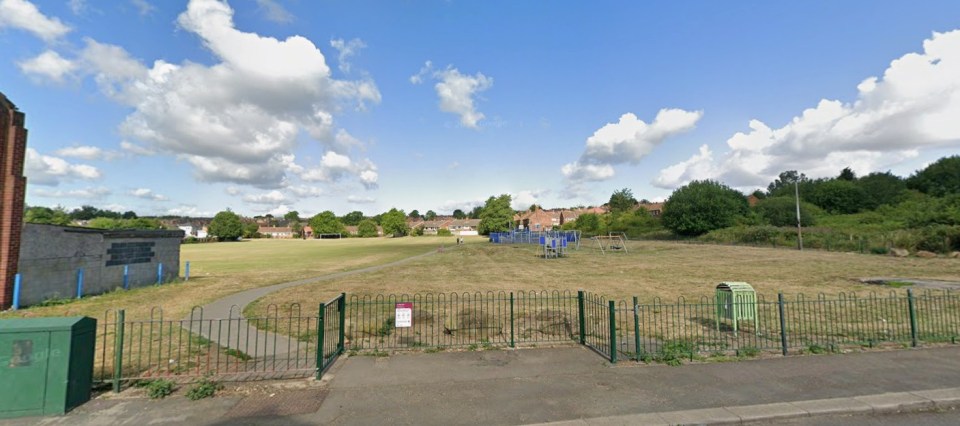 Outdoor view of a dry, grassy field with a playground on the right side and buildings in the background.