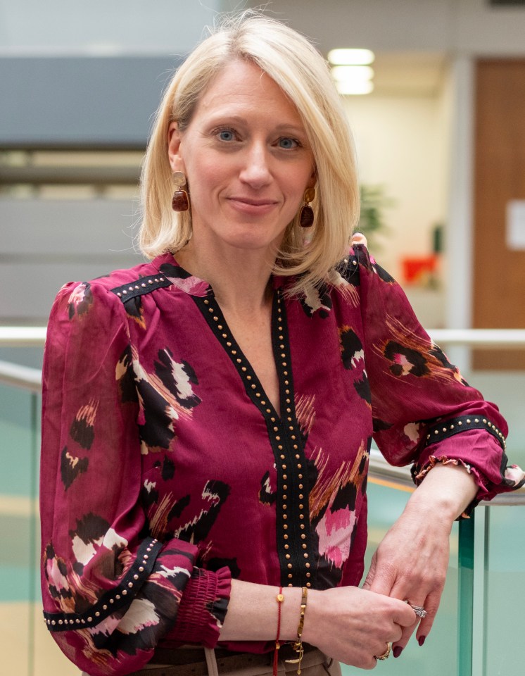 A blonde woman in a deep red blouse with black and gold stud details leans on a glass railing.