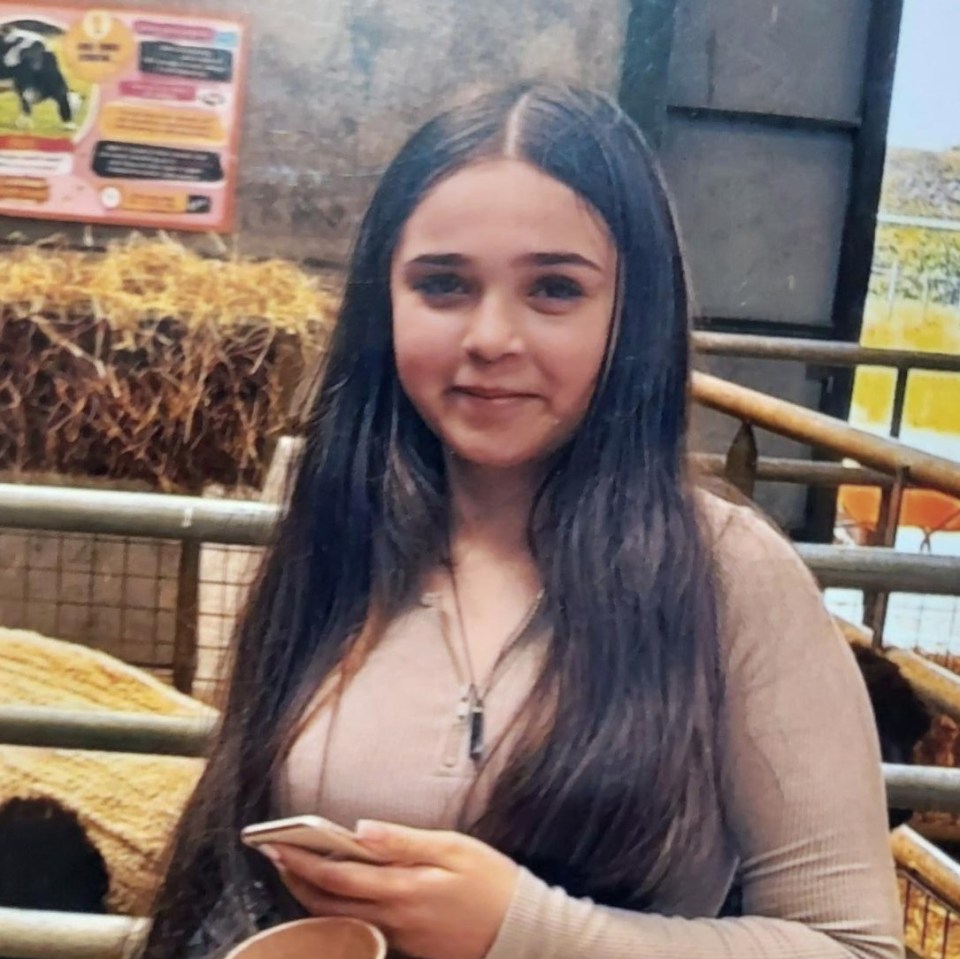 Kerry, a young girl with long dark hair, wearing a beige long-sleeved top and a necklace, holding a phone.