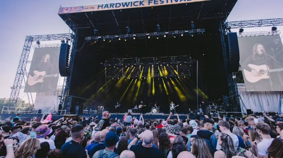 Crowd watches a band perform on stage at Hardwick Festival, with large screens displaying the lead singer.