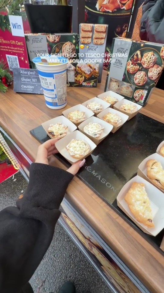 A person's hand reaches for a mini pie at a Tesco market stall, with other pies, cream, and "Tesco Finest" packaging visible.