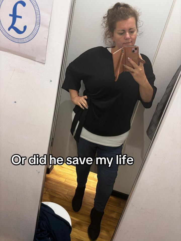 A woman in a black top and blue jeans takes a mirror selfie in a fitting room.