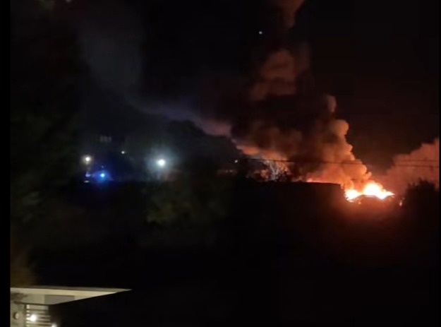 Nighttime photo showing a large fire engulfing a car dealership with thick smoke rising.