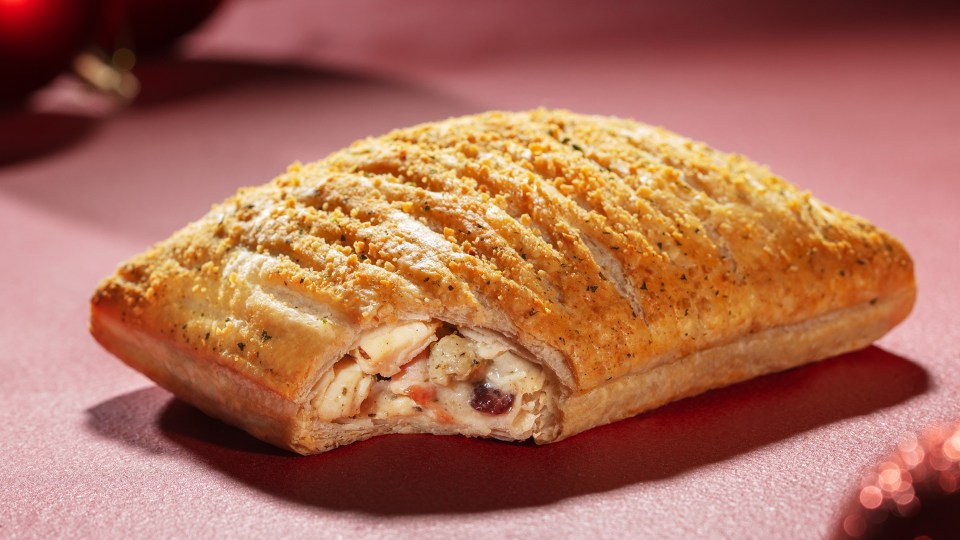 A Greggs Festive Bake on a red surface, with a bite taken out of it revealing the filling.