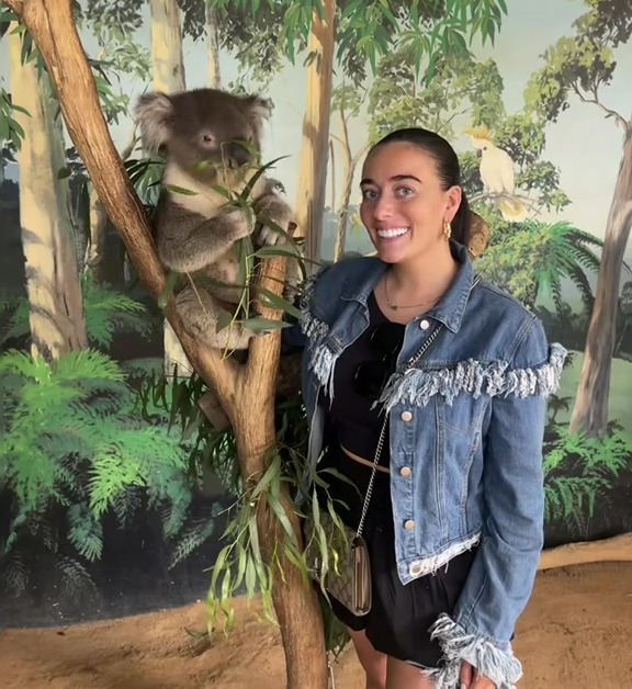 A young woman smiles next to a koala in a tree.