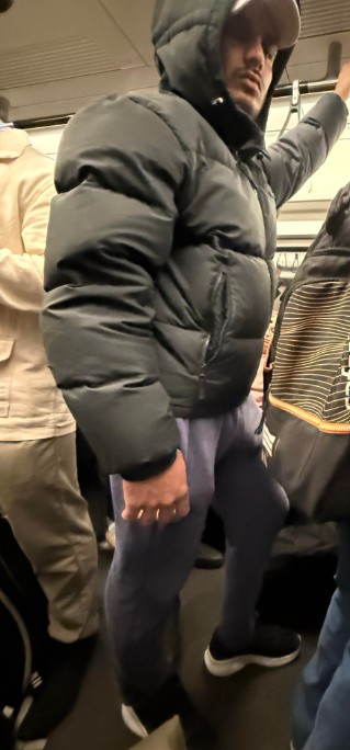 A man in a black puffer jacket, grey cap, and blue sweatpants standing on a crowded public transport.