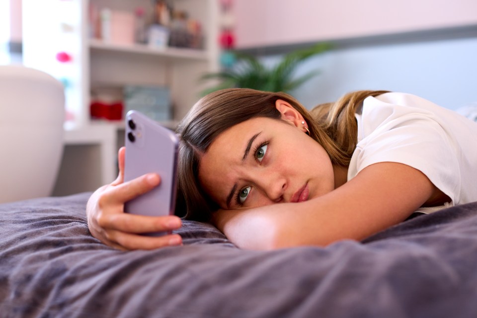 A depressed teenage girl lying on her bed, looking at her mobile phone.
