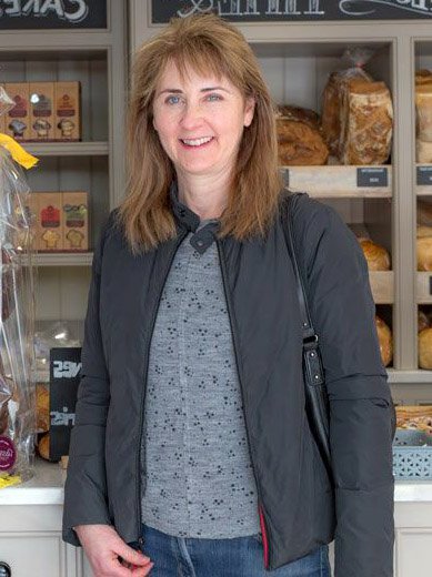 A smiling woman with light brown hair and a dark jacket in a bakery.