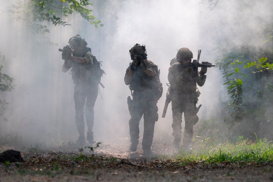 Three armed soldiers in military uniforms walking through heavy smoke in a forest.