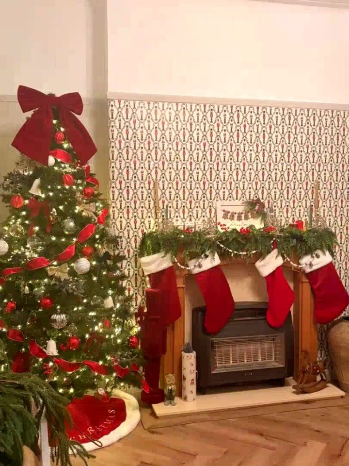 A brightly lit Christmas tree next to a fireplace with four red stockings hanging from the mantelpiece.