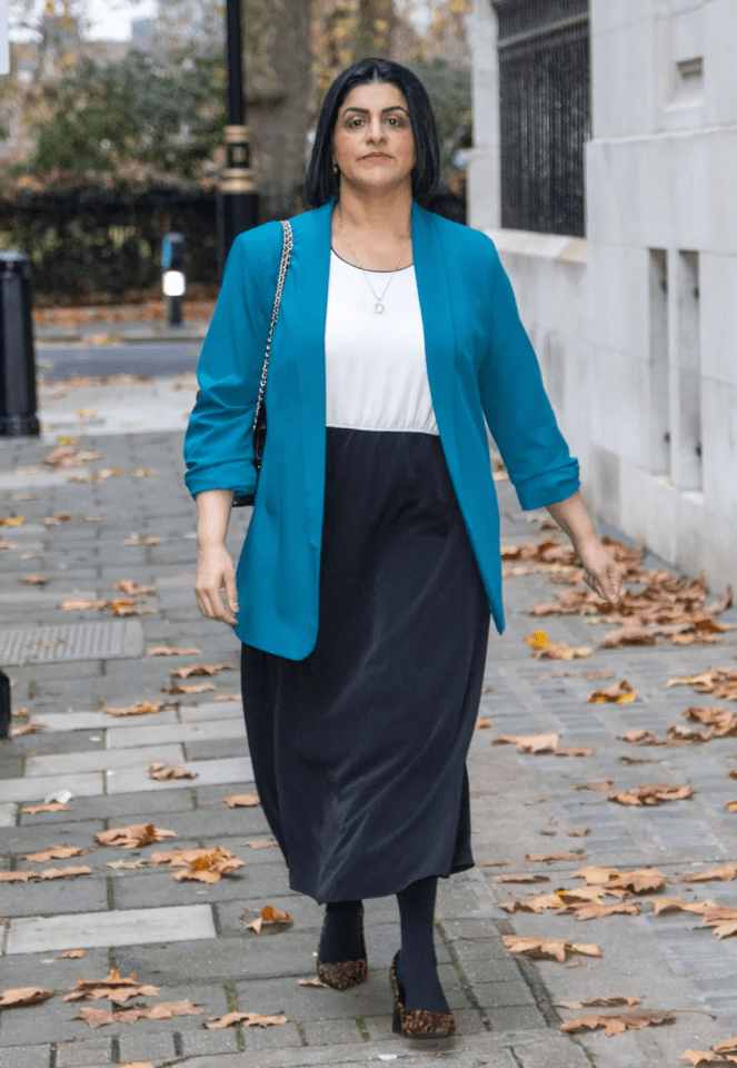 A woman with dark hair wearing a white top, a dark skirt, a teal blazer, and leopard print shoes walks on a sidewalk covered with autumn leaves.