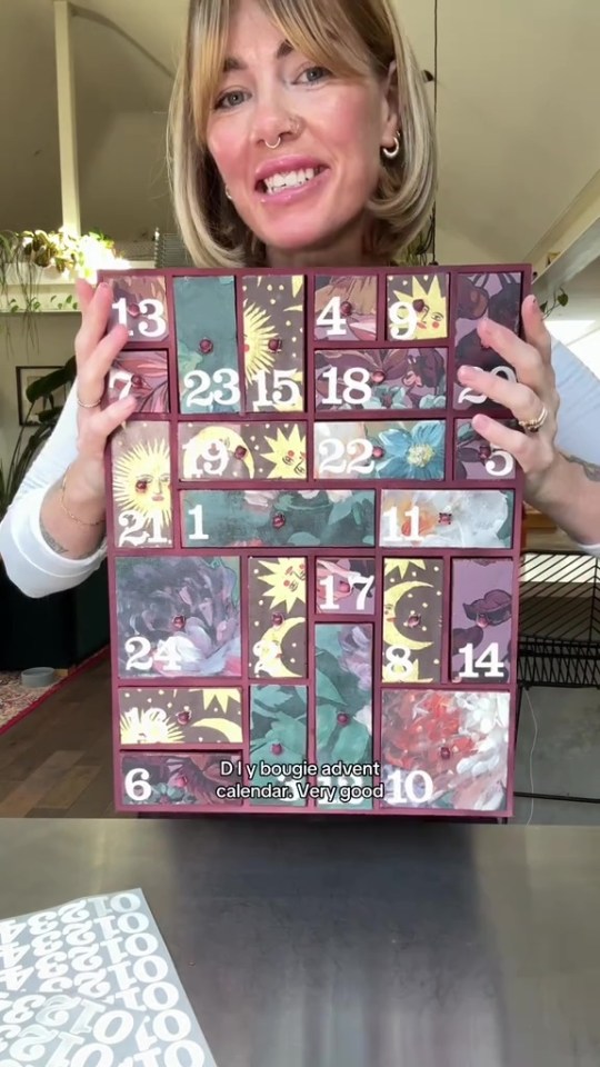 A woman holding a DIY advent calendar made from painted wooden drawers with numbers and decorative papers.