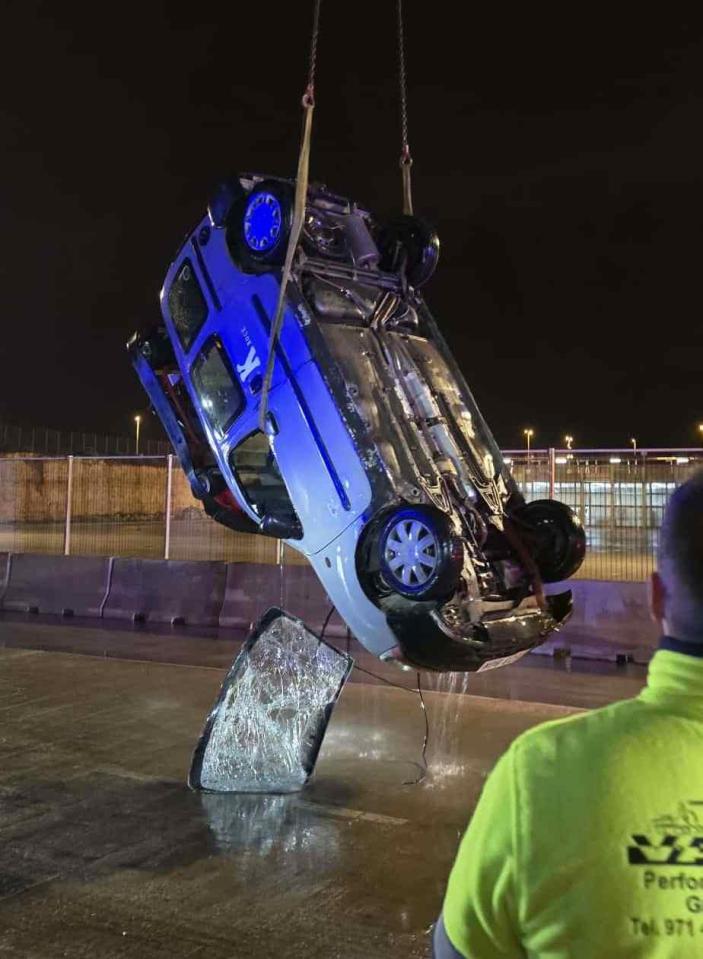 A blue and white car being lifted upside down by a crane from the water at night, with a person in a safety vest watching.
