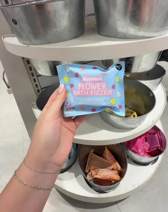A person holds a blue "Bubbles Flower Bath Fizzer" in a store display with metal bins.