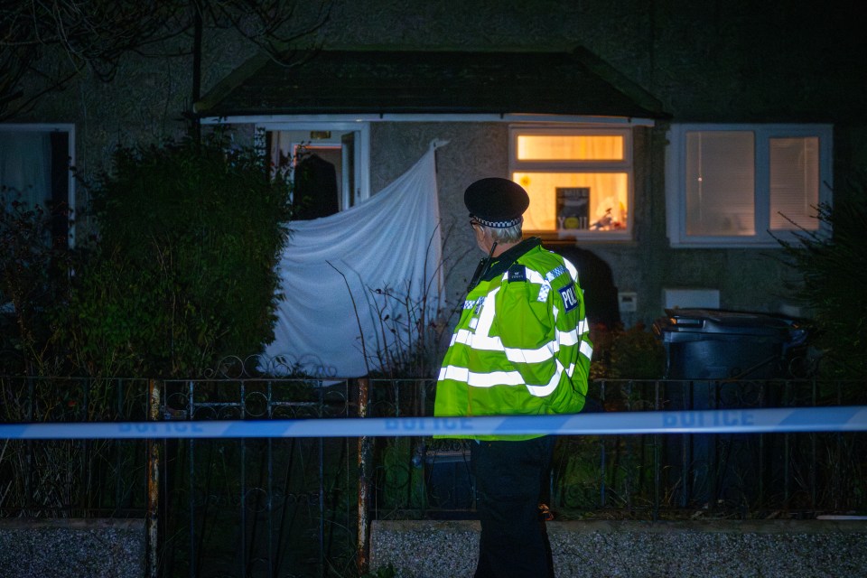 A police officer in a high-visibility vest stands behind police tape in front of a house with a white sheet covering a broken window.