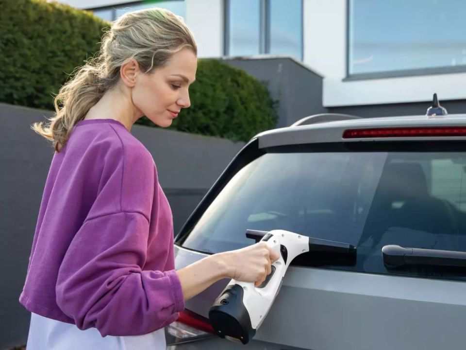 A woman cleans a car window with a white and black window vacuum cleaner.