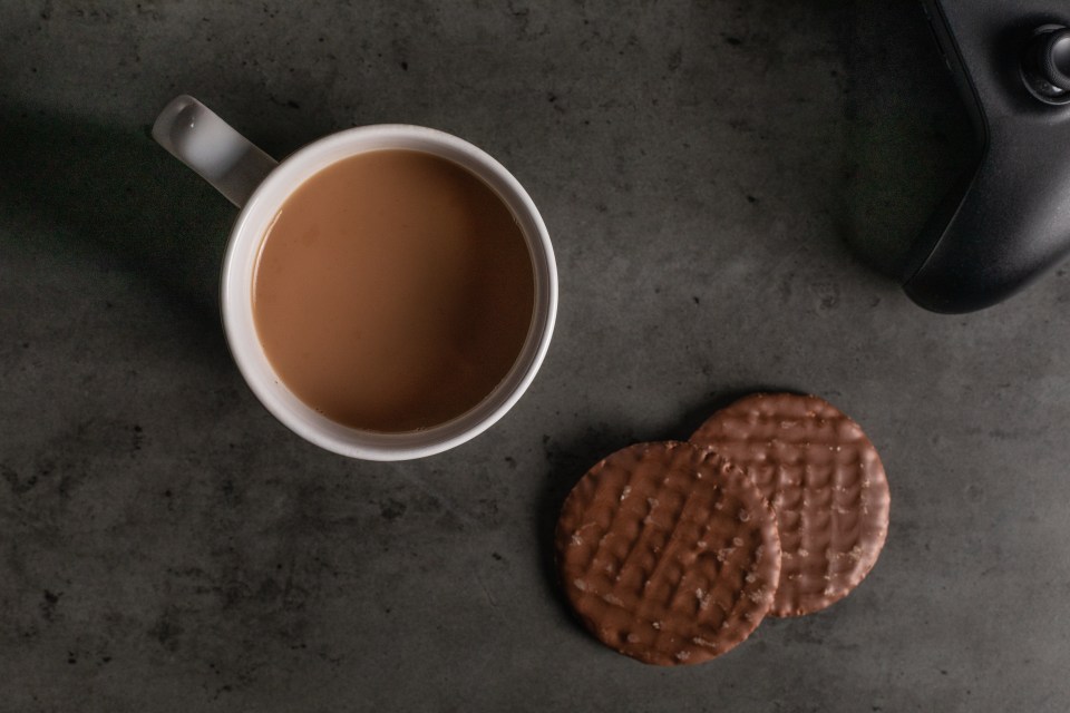 White coffee mug, chocolate digestive biscuits, and game controller on a dark textured table.