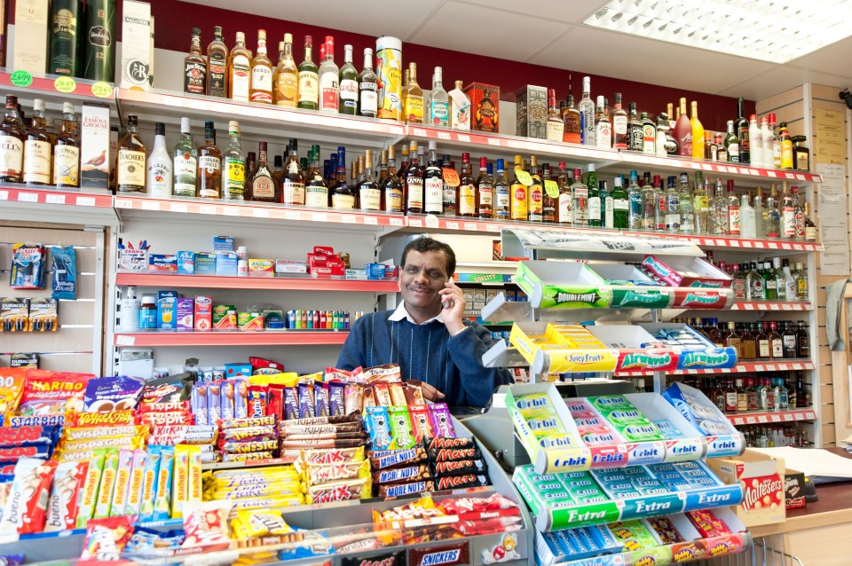 Indian off-licence owner in his London corner shop, surrounded by shelves of alcoholic beverages, candies, and other products, while talking on the phone.