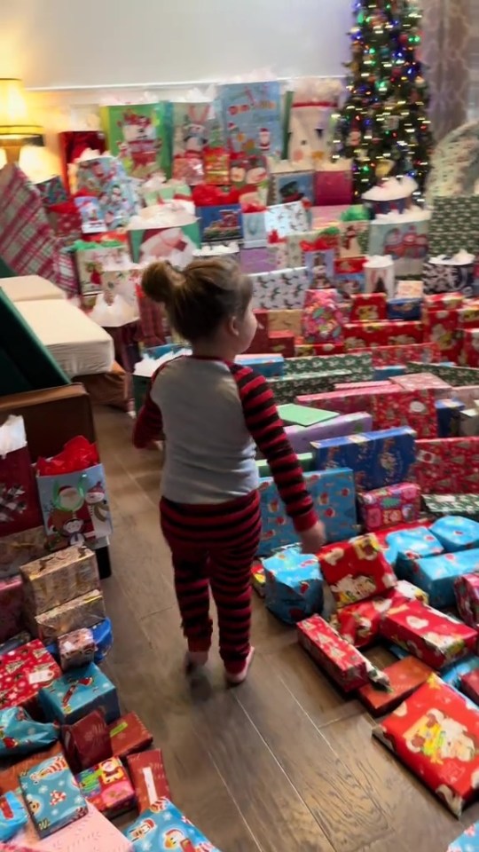 A child in red and black striped pajamas stands in a room filled with wrapped Christmas presents, with a Christmas tree in the background.