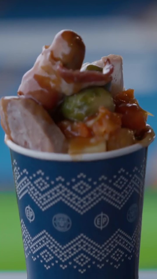 A blue cup with a white festive pattern filled with a Christmas dinner, including turkey, gravy, a pig in a blanket, and sprouts.