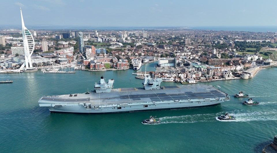 The HMS Prince of Wales aircraft carrier returns to Portsmouth Harbour, escorted by several tugboats, with the city and Spinnaker Tower in the background.