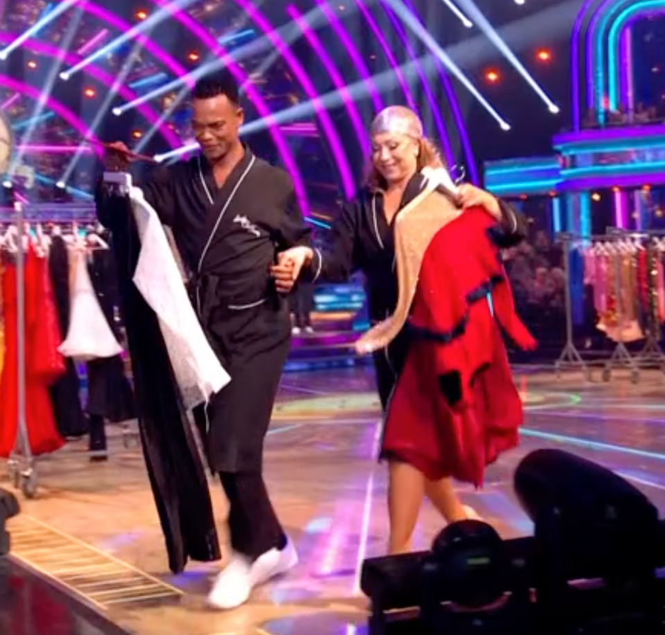 Two people in black robes carrying clothing, walking together on a stage.