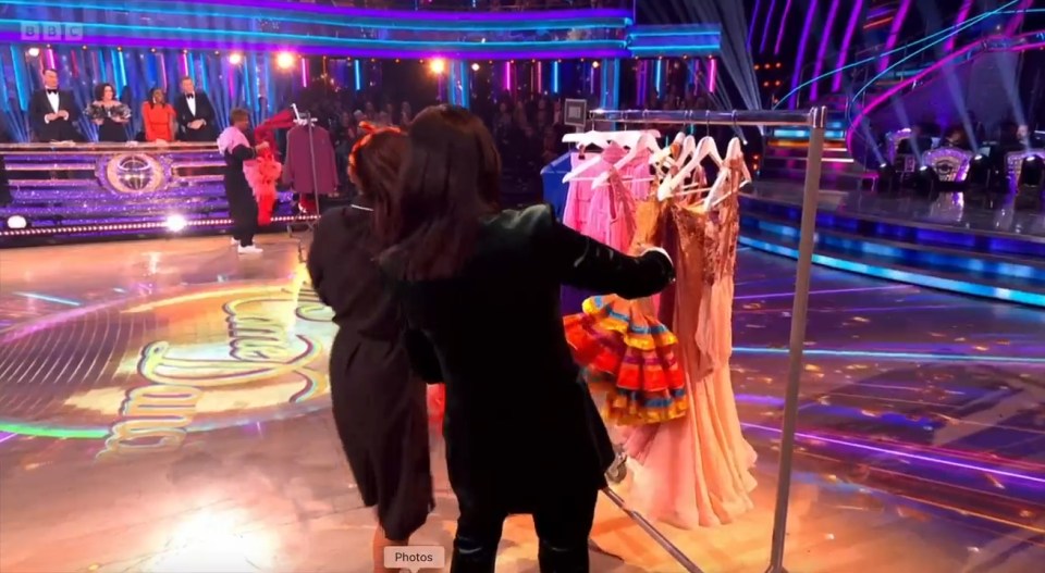 A live dance show with contestants holding various dance costumes, with judges in the background.