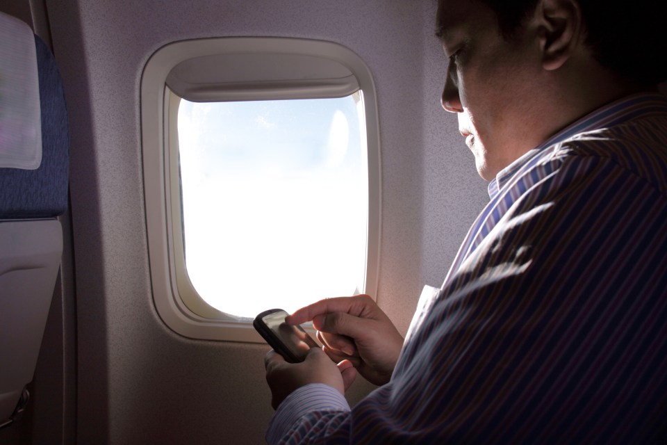 A man uses his phone while sitting next to an airplane window.