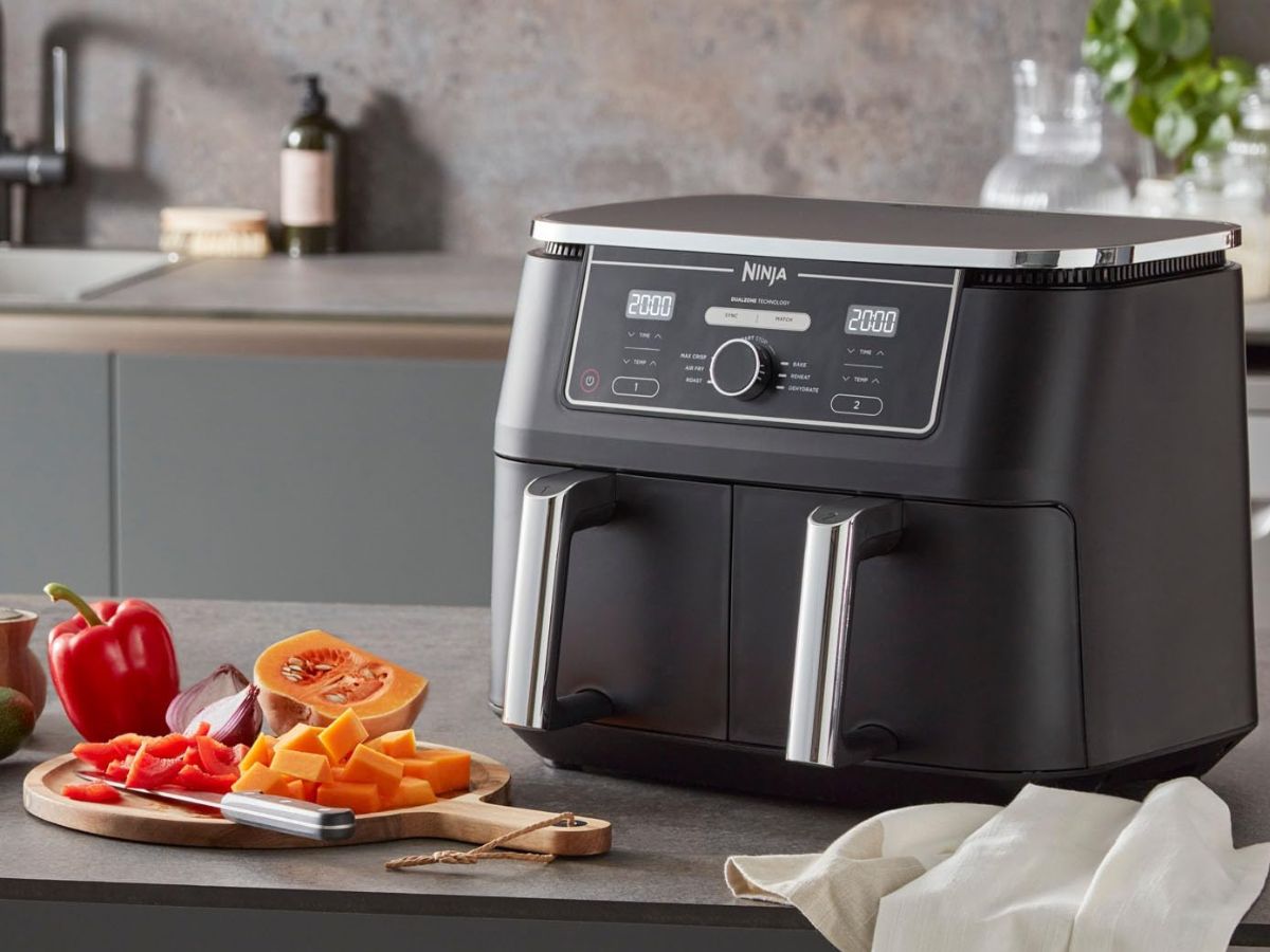 A black Ninja air fryer with dual compartments and digital display on a kitchen counter with chopped vegetables.