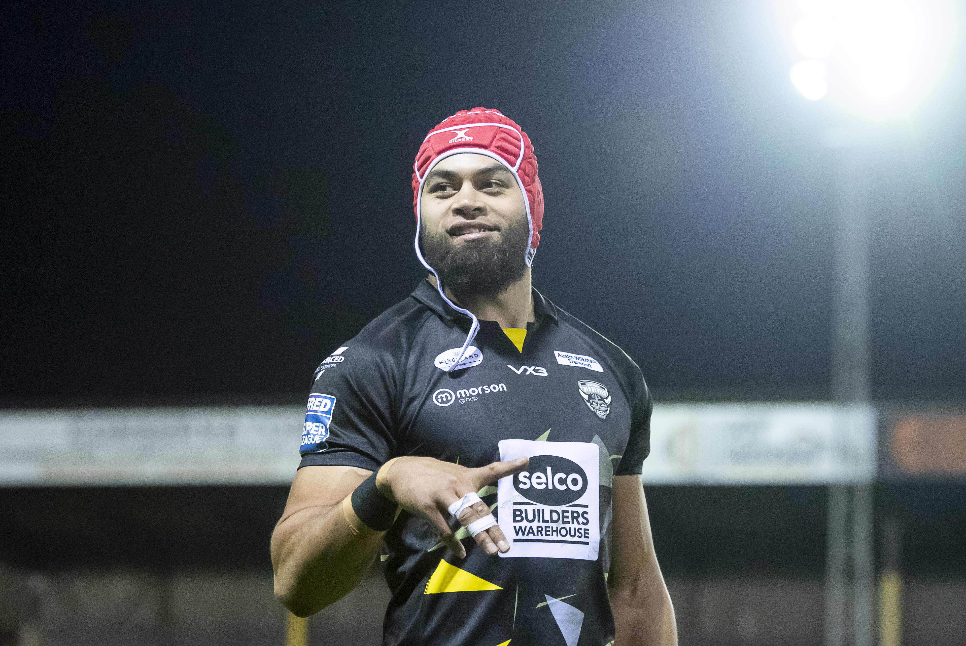 A rugby player in a red scrum cap and a black jersey with sponsor logos, pointing to a "Selco Builders Warehouse" logo on his chest.