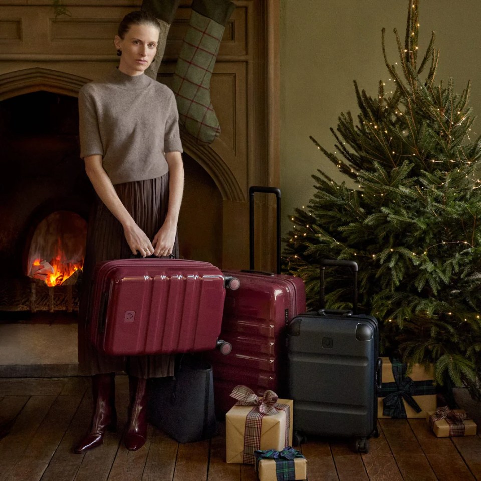 A woman holding a burgundy carry-on suitcase, standing next to a burgundy check-in suitcase, a black carry-on suitcase, wrapped presents, and a Christmas tree by a fireplace.