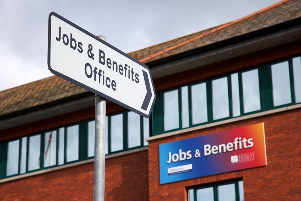 A road sign pointing to a "Jobs & Benefits Office" in front of a brick building with a "Jobs & Benefits" sign.