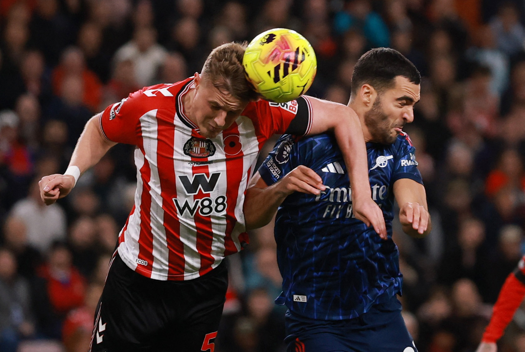Two soccer players in red/white and blue jerseys heading a ball.