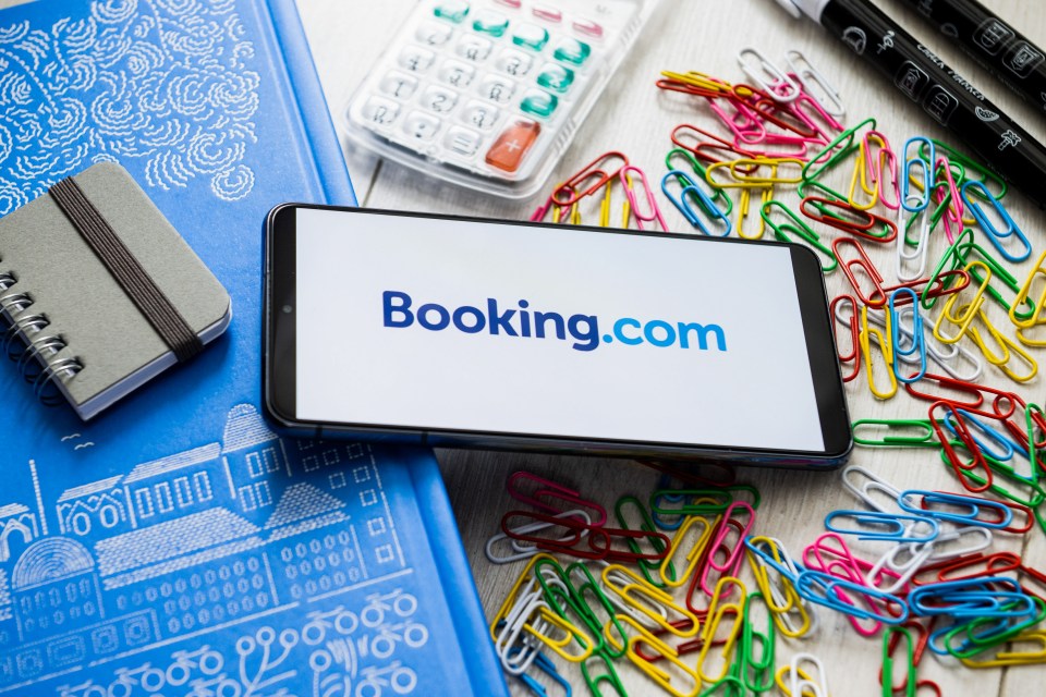 Illustration of a smartphone displaying the Booking.com logo, surrounded by colorful paperclips, a calculator, pens, and notebooks.
