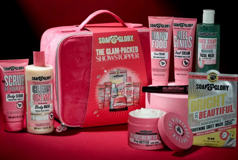 A collection of Soap & Glory products, including body scrub, body wash, hand cream, foot cream, facial wash, body butter, and a sheet mask, arranged around a pink Soap & Glory cosmetic case.