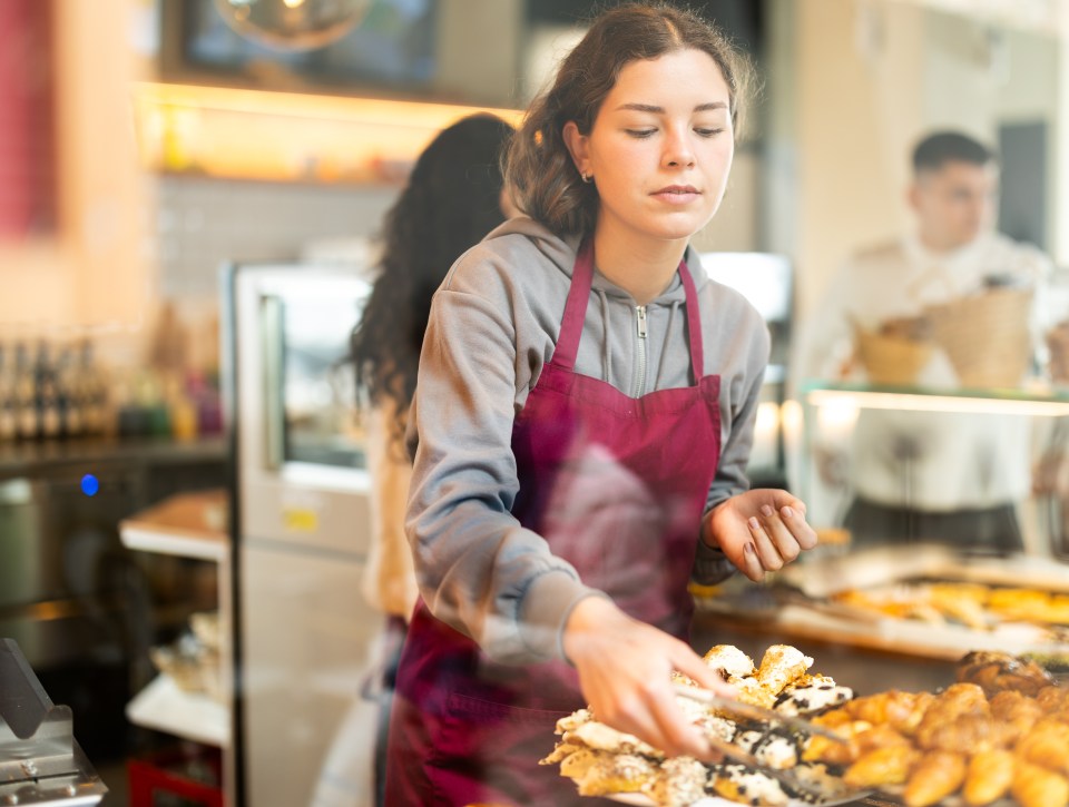A young female baker arranging pastries in a bakery display case.