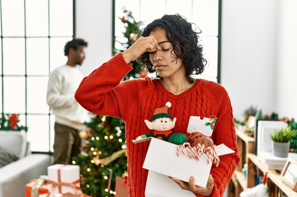 Young Hispanic woman in a red sweater rubbing her nose and eyes, holding a box of Christmas decorations, indicating fatigue and headache, while a man stands blurry in the background near a Christmas tree.