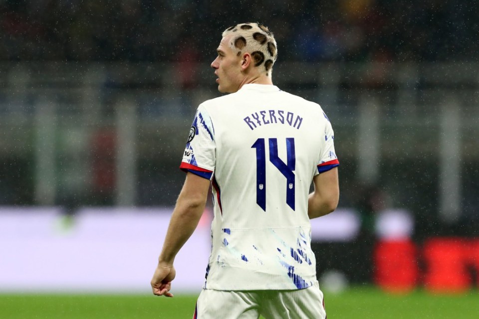 A soccer player in a white jersey with a leopard print haircut, seen from behind, on a rainy field.