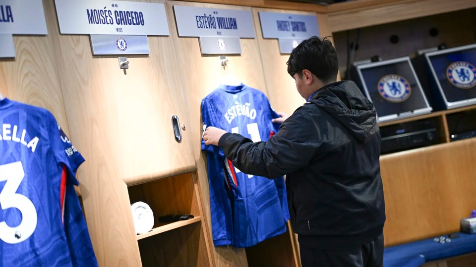 A young boy in a black jacket touching a blue Chelsea F.C. jersey with "ESTEVÃO" and number "11" on the back in a locker room setting.