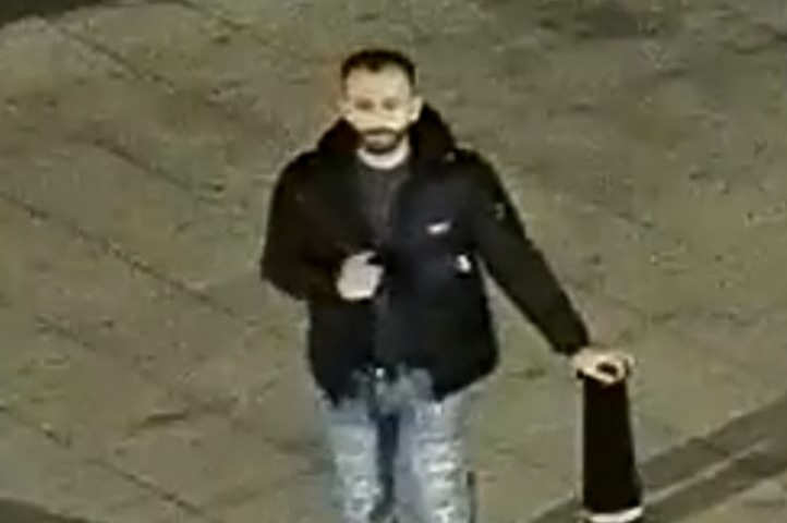 CCTV image of a man with a beard and dark hair wearing a black jacket and light-colored jeans, standing on a sidewalk next to a black bollard.