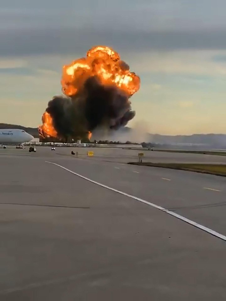 A large explosion with fire and black smoke at an airport, with a commercial aircraft on the tarmac in the foreground.