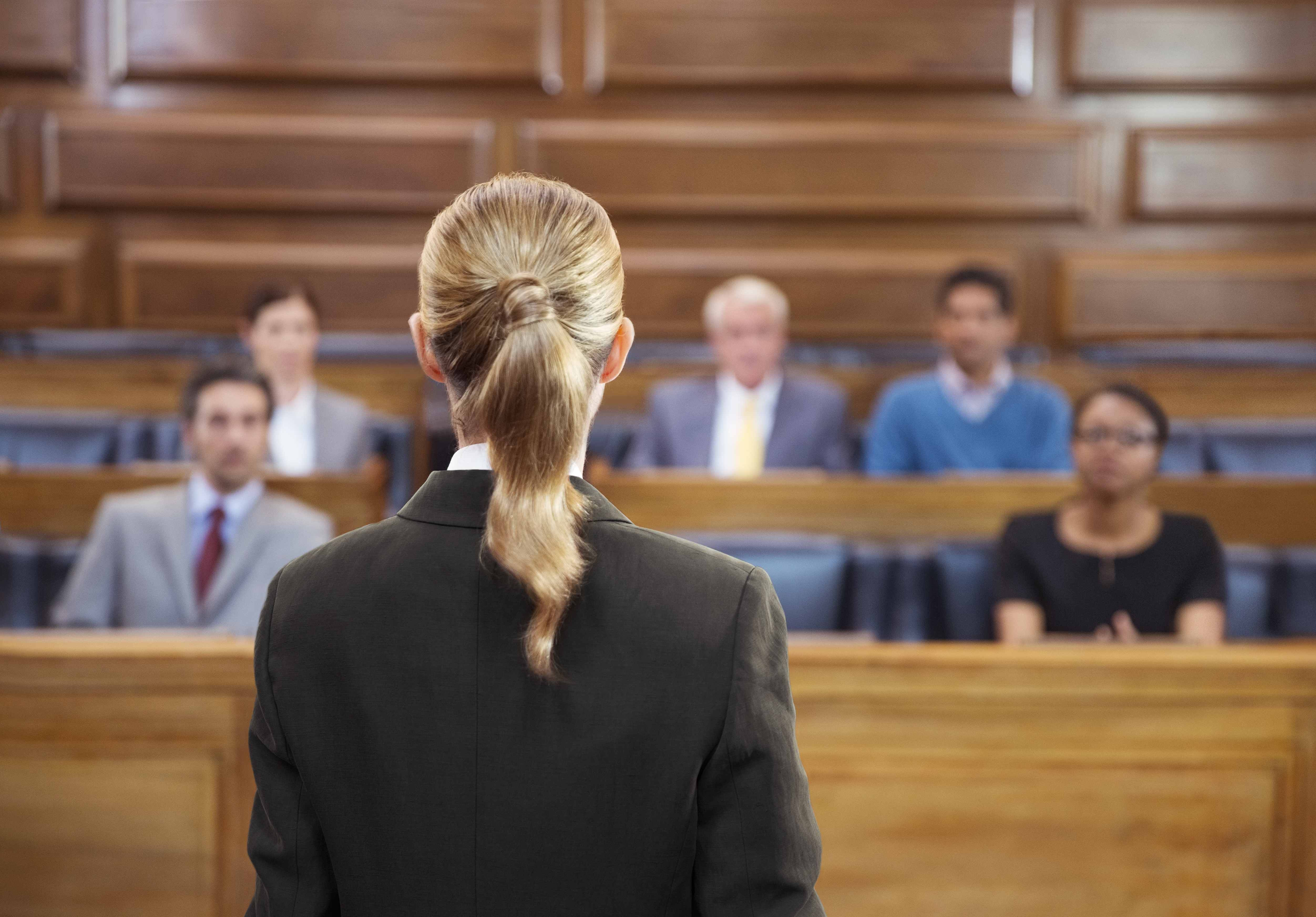 A blonde female lawyer from behind, pleading her case to a jury in a courtroom.