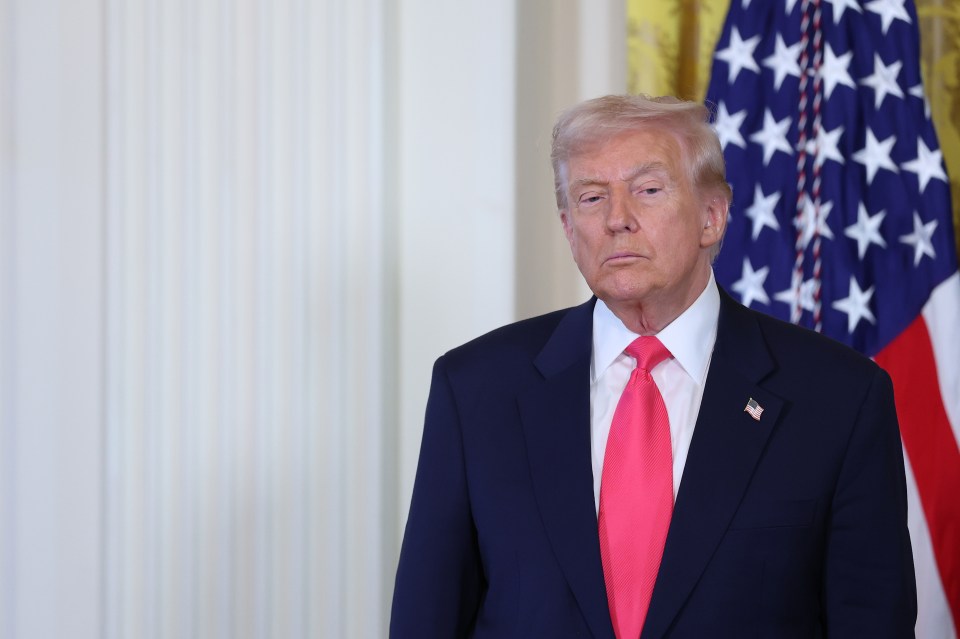 President Donald Trump in a navy suit with a red tie, an American flag pin, and an American flag in the background.