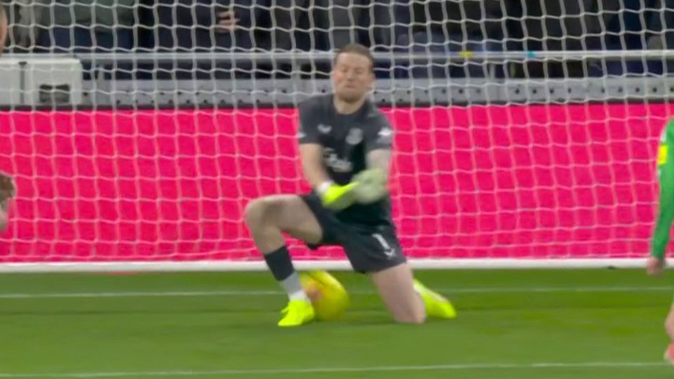 Jordan Pickford, a male soccer player, kneeling on the field in front of a goal with a ball under his knee.